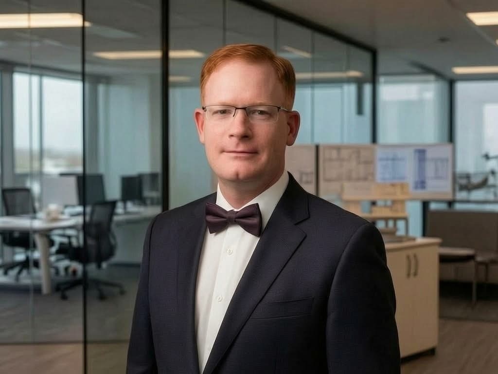 Red-haired man wearing glasses, a suit, and a bow tie in a modern office setting.